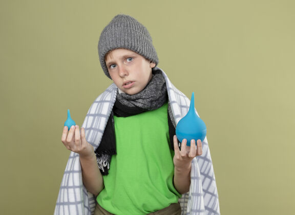 生病生病的小男孩戴着暖和的帽子 围着围巾 裹着毯子 拿着灌肠剂 站在轻薄的墙上发愁暖暖的毯子小