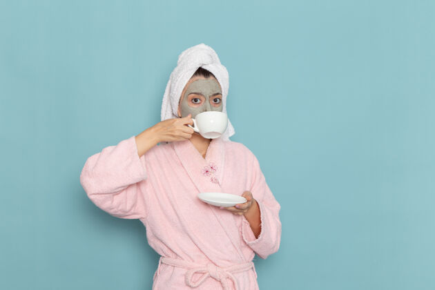 面部正面图身着粉色浴袍的年轻女性淋浴后在浅蓝色墙壁上喝咖啡美丽的清水自理淋浴浅蓝色清洁自我护理