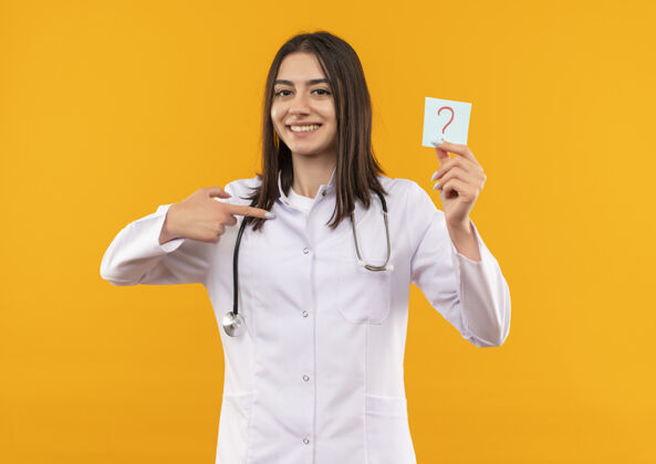 办公室年轻的女医生穿着白大褂 脖子上戴着听诊器 手里拿着带问号的提醒纸 手指指着提醒纸 微笑着看着站在橙色墙上的前面标记医院指着