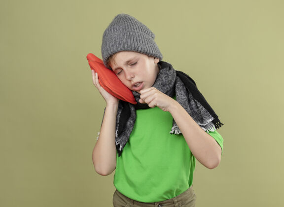 不适生病的小男孩穿着绿色t恤 戴着暖和的围巾和帽子 感觉不舒服 拿着水瓶取暖 把头靠在上面 站在轻墙上咳嗽瓶子穿头