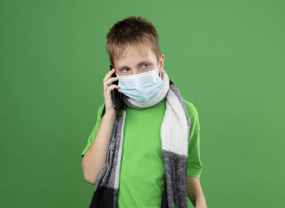 面部生病的小男孩穿着绿色t恤 脖子上围着暖和的围巾 戴着面罩 站在绿色的墙上讲着手机小电话谈话