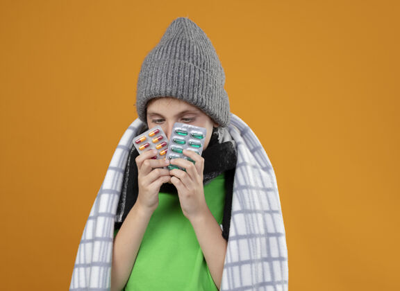 感觉生病的小男孩戴着暖和的帽子 围着围巾 裹着毯子 站在橘色的墙上发烧 吃着避孕药 感觉不舒服帽子疾病站着