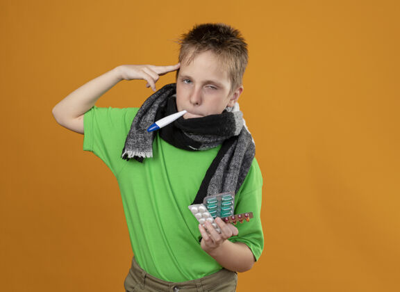 小生病的小男孩穿着绿色t恤 脖子上围着暖和的围巾 嘴里叼着体温计 手里拿着药片 站在橘色的墙上看着自己的太阳穴 看上去很不舒服围巾站着看