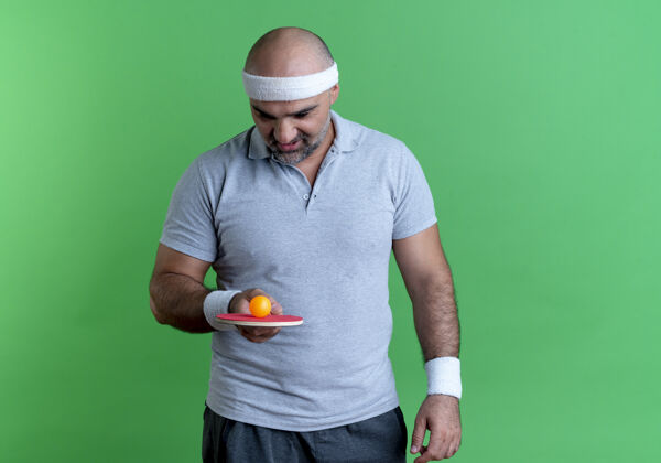 运动戴着头巾的成熟运动型男子拿着球拍和乒乓球站在绿色的墙上饶有兴趣地看着它姿势运动员男