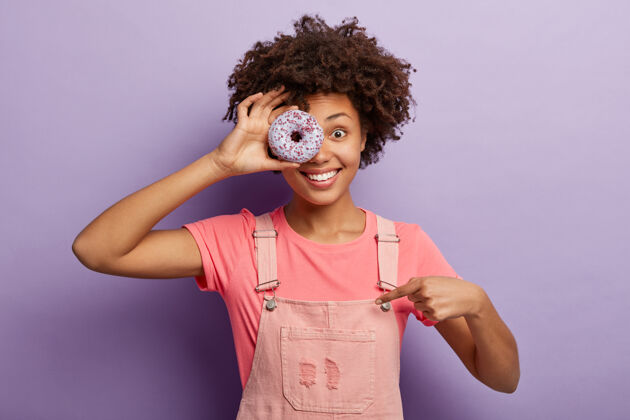女性饥饿的微笑女孩用甜甜的甜甜圈遮住一只眼睛 有不健康的营养 指着粉色的工作服 打破饮食美味工作服封面