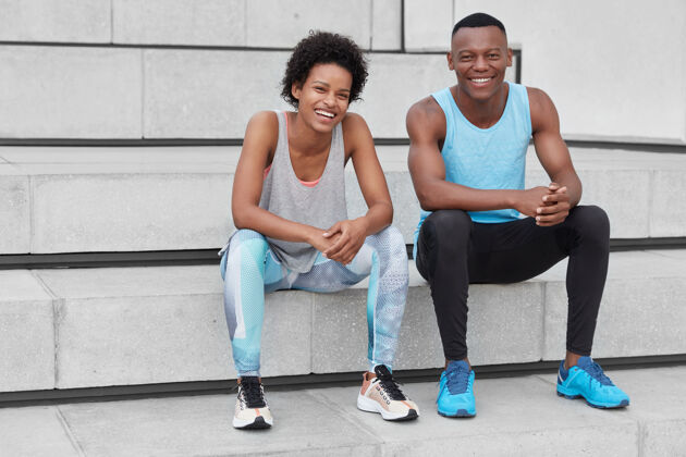 满意镜头中的非洲裔美国人轻松无忧无虑 表情积极 穿着运动服 坐在楼梯上 愉快地谈论着体育新闻友谊 训练快乐运动楼梯