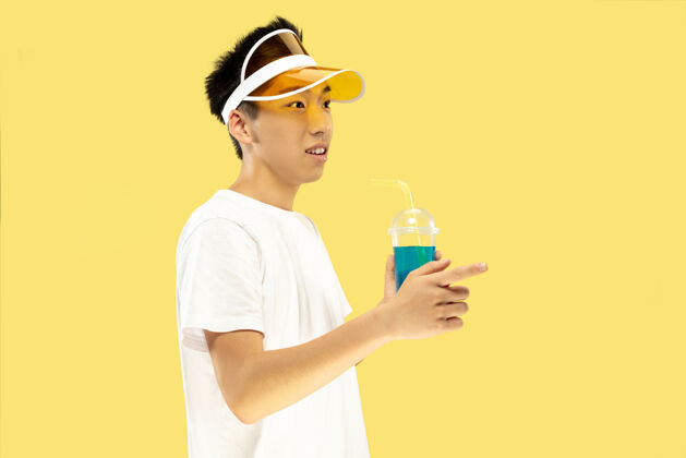 成功黄色工作室背景上的韩国年轻人半身像穿着白衬衫 戴着黄色帽子的男模喝鸡尾酒人类情感的概念 表达 夏天 假期 周末惊奇周末面部