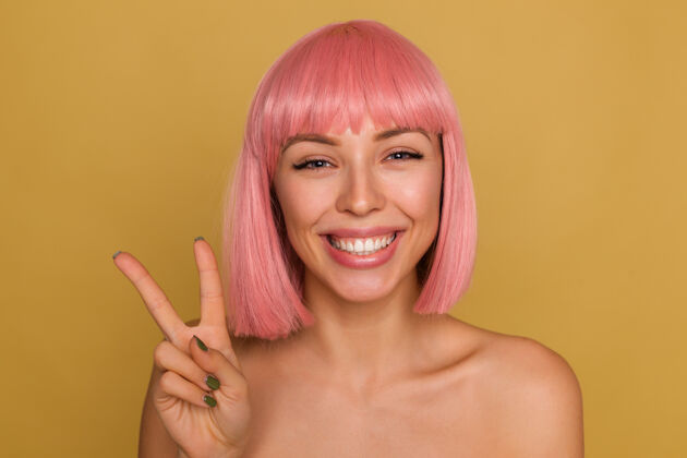 年轻快乐的年轻迷人的女士 粉色时髦的bob发型 露出洁白完美的牙齿 面带微笑 举手投足 手举胜利标志 隔着芥末墙20岁特写鲍勃