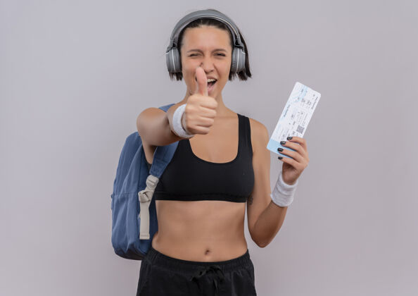 举行身穿运动服 头戴耳机 背着背包 拿着机票 微笑着竖起大拇指的年轻健身女士站在白墙上微笑健身运动