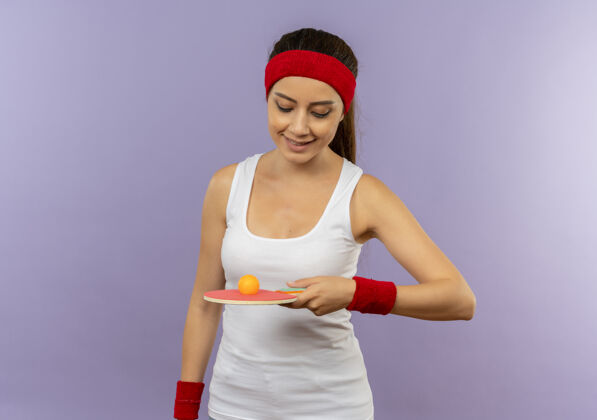 人身穿运动服的年轻健身女 头戴钢圈 手里拿着乒乓球和球拍 站在灰色的墙上 面带微笑地看着它人年轻运动员