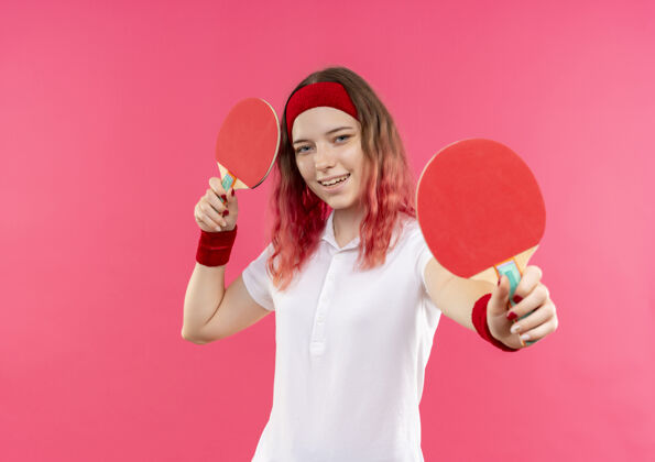 年轻戴着头巾 拿着两个乒乓球拍的年轻运动女郎站在粉红色的墙上 面带幸福的笑容桌子姿势女人
