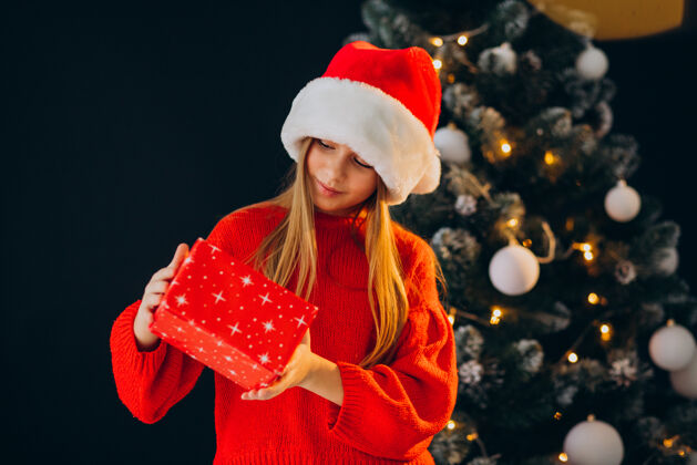 礼物可爱的少女戴着圣诞树旁的红色圣诞帽看可爱庆祝