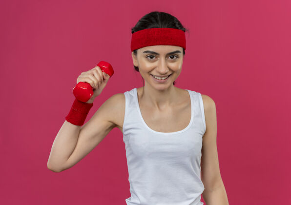 健身身穿运动服 头箍 举着哑铃 面带微笑 自信地站在粉色墙壁上锻炼身体的年轻健身女士举起来人摆姿势