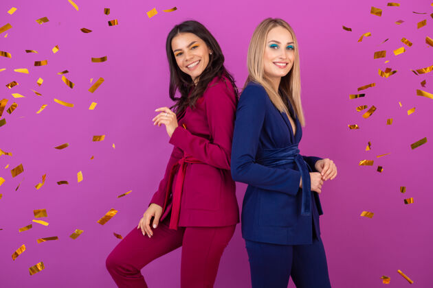 快乐两位魅力四射的女士在紫罗兰色的墙上欢庆新年 穿着紫色和蓝色的时尚多彩的晚礼服 朋友们在一起玩得很开心 时尚潮流 金色的五彩纸屑派对气氛欢快人五彩纸屑