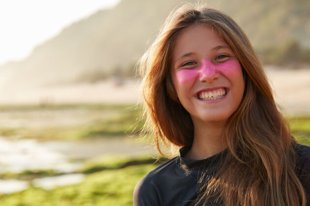 姿势乐观的年轻快乐的欧洲女人 带着牙齿般的笑容 脸上戴着能阻挡阳光的锌防护面具 穿着潜水服冲浪 在户外对着模糊的海岸线墙摆姿势海滩冲浪者白种人