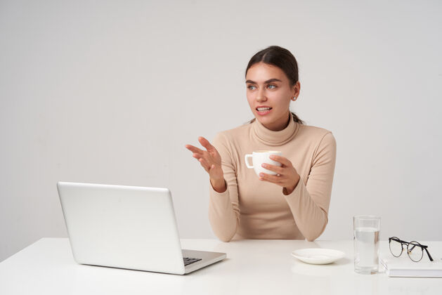 模特一位年轻漂亮的黑发女商人正在和同事们开会 她穿着米黄色的马球衫坐在白色的墙上 手里捧着一杯茶 举起手来女人情感笔记本电脑