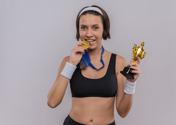 金牌一位身穿运动服 脖子上挂着金牌的年轻健身女士高高兴兴地拿着奖杯 站在白墙上积极地咬着奖牌奖杯女性周围