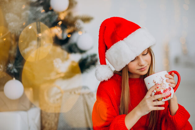 圣诞节在圣诞树旁喝可可的女孩青少年节日圣诞帽