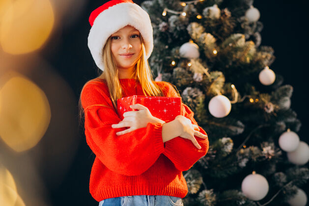 灯可爱的少女戴着圣诞树旁的红色圣诞帽传统看红帽子