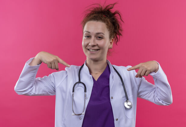 听诊器年轻的女医生穿着白大褂 用听诊器指着自己满意地站在粉红色的墙上医生她自己自我满足
