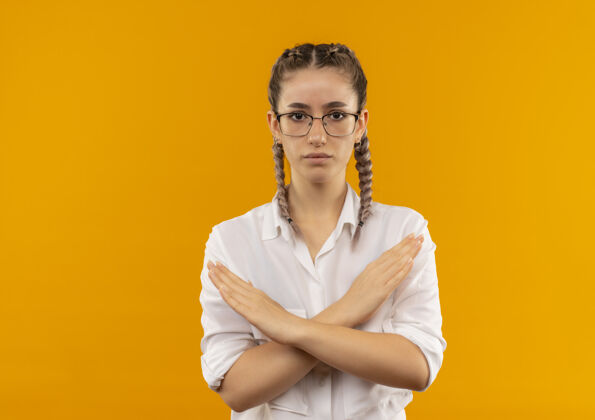 手势戴着眼镜 梳着辫子 穿着白衬衫的年轻女学生正脸交叉双臂站在橘色的墙上做停车标志脸公民眼镜