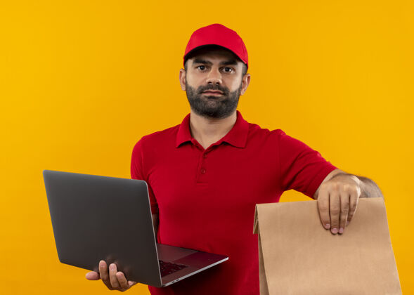 站着有胡子的送货员穿着红色制服 戴着帽子 手里拿着纸包和笔记本电脑 严肃的脸站在橙色的墙上脸拿着包裹