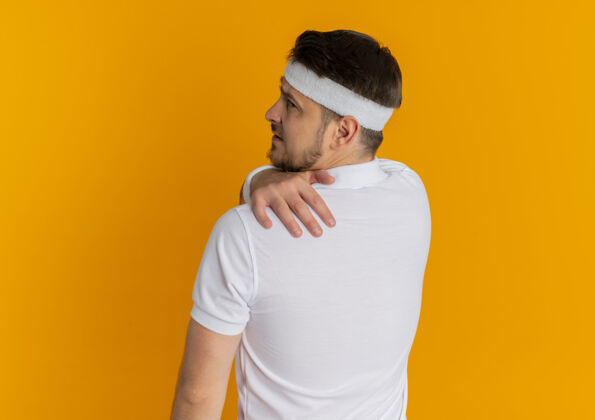 站立身穿白衬衫 头戴头巾 背靠肩站立的年轻健身男子在橘红色的墙壁上感到疼痛教练头带背部