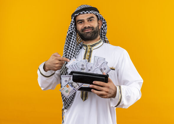 穿身着传统服饰的阿拉伯商人手拿钱包 用手指指着现金 自信地站在橙色的墙上商务人士手指现金