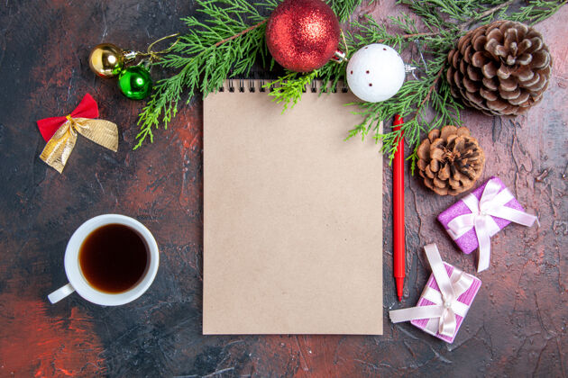 顶部顶视图红色钢笔笔记本松树树枝圣诞树球玩具和礼物一杯茶在暗红色的表面背景笔记本茶