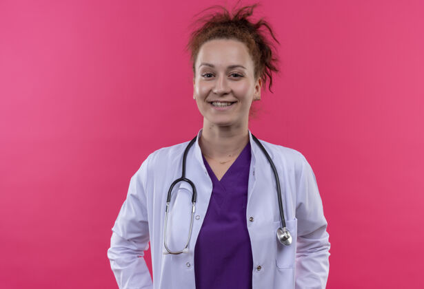 女士身穿白大褂 手持听诊器 面带微笑的年轻女医生站在粉红色的墙上听诊器微笑站着