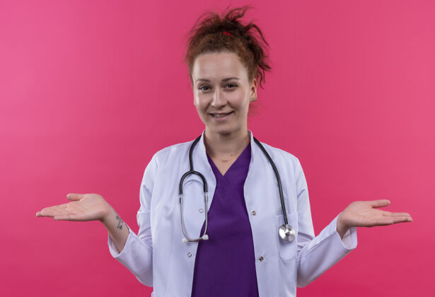 摊开年轻的女医生身穿白色外套 手持听诊器 微笑着张开双臂站在粉红色的墙上女人听诊器微笑