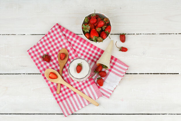视野俯视图：一碗草莓放在红色方格毛巾上 用木勺 一个草莓筒和一碗酸奶放在白色木板上水平吃桌子夏天