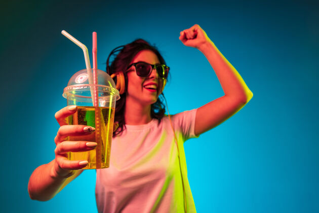 霓虹灯快乐的年轻女子戴着耳机在时髦的蓝色霓虹灯工作室里跳舞和微笑太阳镜度假村饮料