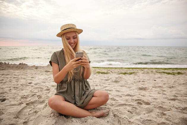 电话迷人的金发女人的肖像 随意的发型 盘腿坐在海边 手里拿着智能手机 愉快地看着屏幕 读着好消息女性长大海