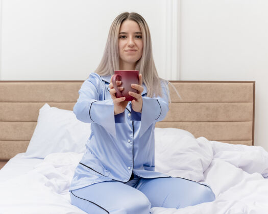 睡衣穿着蓝色睡衣的年轻美女坐在床上 端着一杯咖啡 看着摄像机 在卧室的室内灯光背景下微笑着漂亮杯子坐着