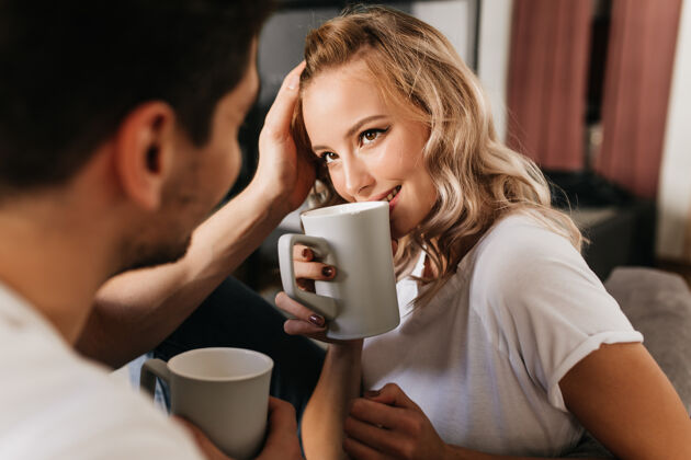 肖像美丽的金发女孩在恋爱中看着她的男朋友 喝着杯子里的咖啡在家里的浪漫情侣的温柔可爱的肖像女性房子看