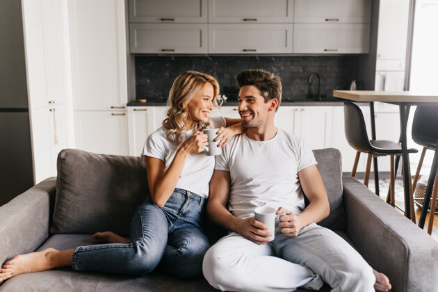 现代相爱的情侣坐在沙发上拿着杯子 面带微笑浪漫的情侣在家里享受早晨看杯子在一起