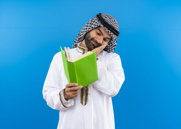阿拉伯语身着传统服饰的阿拉伯商人手持一本打开的书 面带微笑地站在蓝色的墙上看着它持有站脸