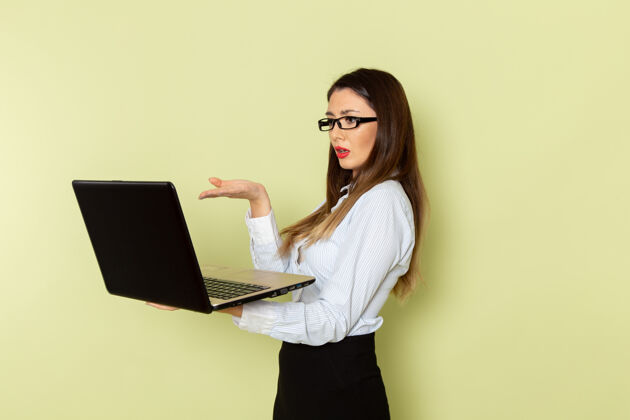 便携式穿着白衬衫和黑裙子的女上班族正拿着笔记本电脑站在绿色的墙上前面女工作