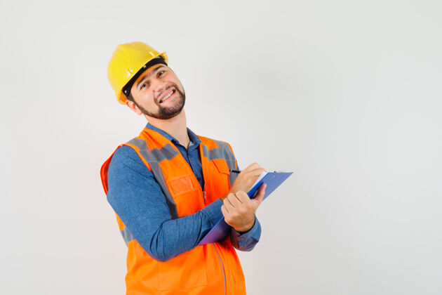 工厂年轻的建筑工人穿着衬衫 背心 戴着头盔在剪贴板上做笔记 看上去很高兴 正前方视图工匠工作工人