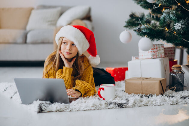 在线可爱的女孩在圣诞树旁用电脑现代快乐的女人红帽子