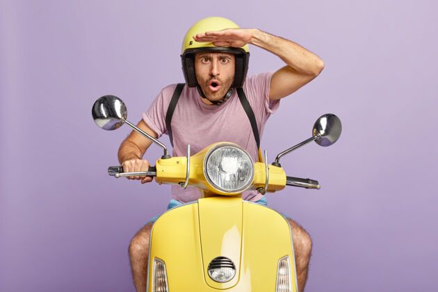 速度惊讶的家伙开着摩托车 把注意力集中在远处 双手放在额头上 戴着黄色头盔和t恤 给顾客发订单 孤立在紫色的墙上震惊的摩托车手看头盔室内