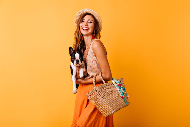 表达戴着夏天帽子的快乐女人抱着法国斗牛犬穿着橙色裙子的欢笑女孩和有趣的小狗合影积极黄色室内