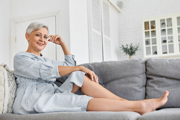 沙发低角度观看时尚优雅成熟的60岁白人女性 短发 在家里放松 坐在灰色沙发上 在她宽敞舒适干净的客厅里 微笑着生活成人舒适