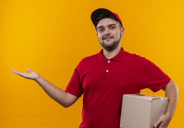 空格年轻的送货员身着红色制服 戴着帽子拿着盒子包裹 用手臂微笑着送礼物盒子演示帽子