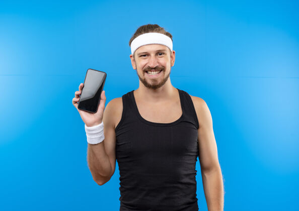 蓝色面带微笑的年轻帅气运动男戴着头带和腕带手持手机隔离在蓝色空间手机手机微笑