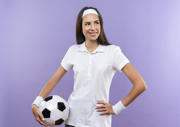 头带微笑着漂亮的运动女孩戴着头带和腕带拿着足球看一边手放在腰上孤立的紫色空间球紫色穿