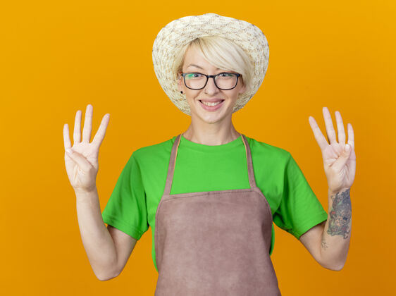 手指一位年轻的园丁 围裙短发 戴着帽子 站在橙色的背景上微笑着 手指向上指着8号围裙向上短裤