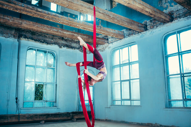 杂技优雅的体操运动员在蓝色的老阁楼上用红色织物进行空中练习柔韧青少年运动