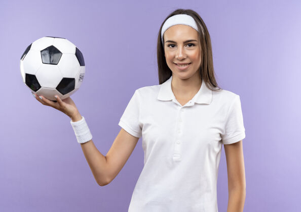 抱着微笑着漂亮的运动女孩戴着头带和手环拿着足球孤立在紫色的空间女孩漂亮足球